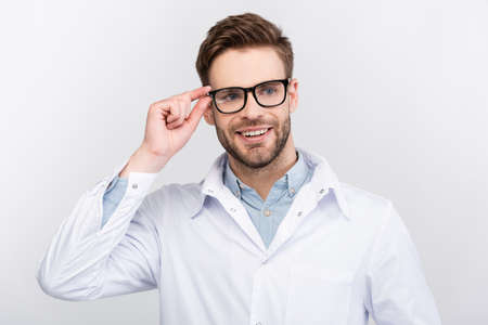 Smiling doctor in white coat touching eyeglasses isolated on gray
