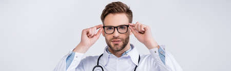 Portrait of young adult doctor holding eyeglasses frame, while looking at camera isolated on white,...