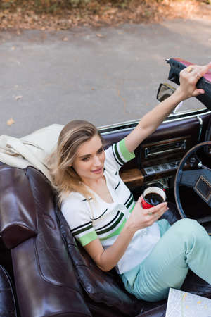 high angle view of young stylish woman holding cup of coffee while sitting in cabriolet near map