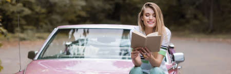 cheerful young woman reading book while sitting on hood of vintage cabriolet on blurred background,...