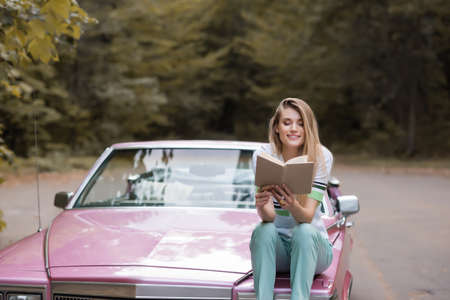 happy young woman reading book while sitting on hood of convertible car on blurred backgroundの写真素材