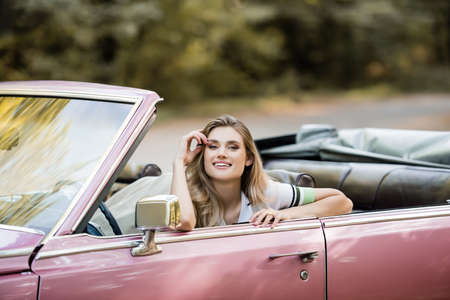 happy young woman holding hand near face while sitting in cabriolet and looking at cameraの写真素材