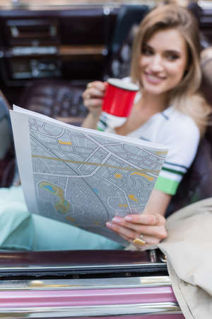 smiling woman holding map and cup of coffee while sitting in cabriolet on blurred background