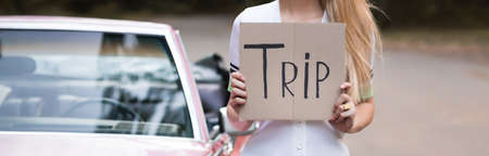 cropped view of woman holding card with trip lettering near vintage cabriolet on blurred background,...