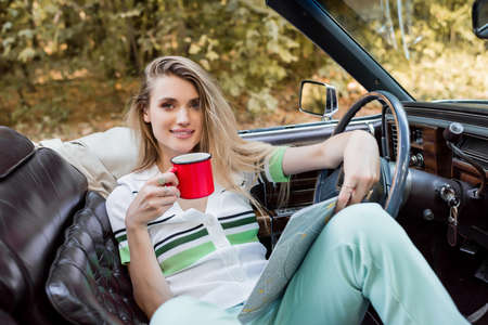 young smiling woman looking at camera while sitting in convertible car with road atlas and cup of coffeeの写真素材