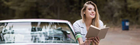 smiling woman sitting on car with book and looking away on blurred background, bannerの写真素材