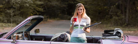 smiling woman looking at camera while holding road atlas and cup of coffee in cabriolet, banner