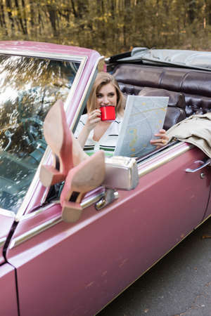 smiling woman drinking coffee and looking at map while putting legs on rear view window on blurred foregroundの写真素材