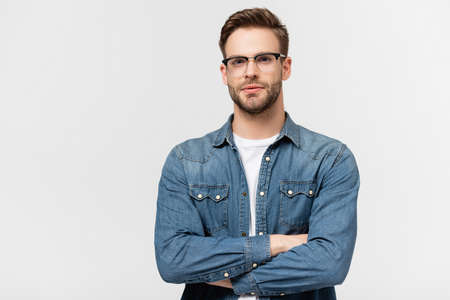 Young man in eyeglasses looking at camera while standing with crossed arms isolated on grayの写真素材