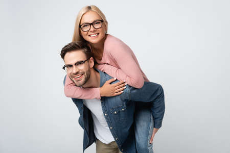 Smiling woman in eyeglasses piggybacking on boyfriend isolated on grayの写真素材