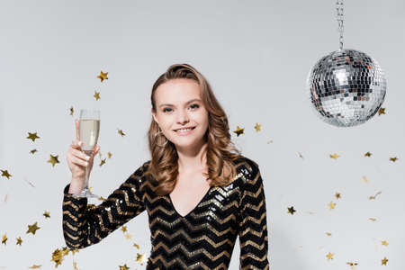 cheerful young woman holding glass of champagne near disco ball and falling confetti on grayの写真素材