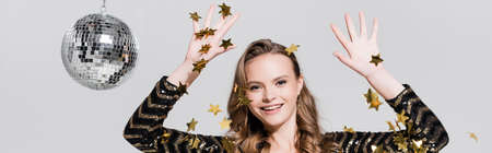 cheerful young woman standing near disco ball and falling confetti on gray, banner