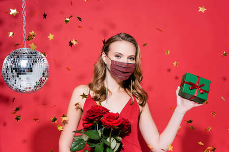 woman in protective mask with rhinestones holding christmas present near confetti and disco ball on...