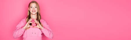 beautiful young woman in hoodie showing heart with hands on pink background, banner