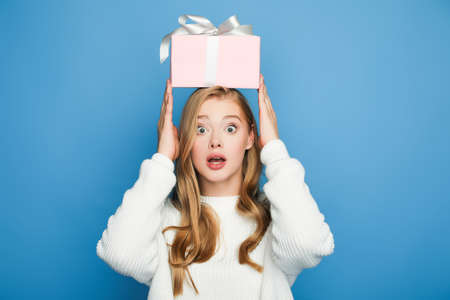 surprised blonde beautiful woman in sweater holding gift box above head isolated on blue
