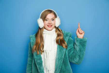 smiling stylish woman in green faux fur coat and ear muffs pointing up isolated on blueの写真素材