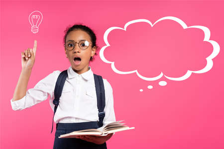 shocked african american schoolgirl in glasses holding book and showing idea gesture near thought...