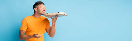 man in t-shirt holding and smelling carton pizza box on blue, banner