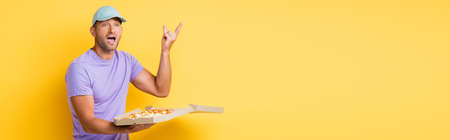 man in blue cap gesturing while holding box with tasty pizza on yellow, bannerの写真素材