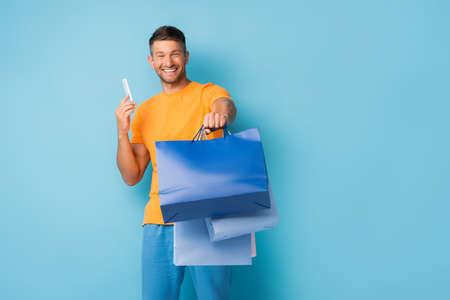 happy man holding shopping bags and smartphone on blue