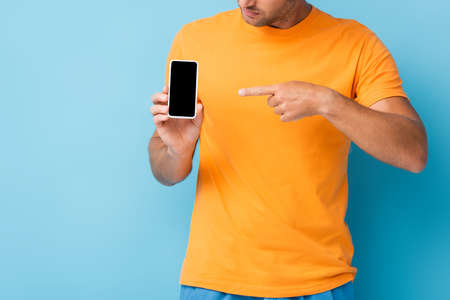 cropped view of man in t-shirt pointing with finger at smartphone with blank screen on blue