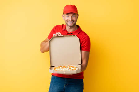 cheerful delivery man in red cap holding box with tasty pizza on yellowの写真素材