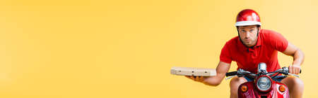 concentrated delivery man in helmet riding red scooter and holding pizza box isolated on yellow, bannerの写真素材