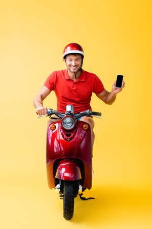 happy delivery man in helmet riding scooter and holding smartphone with blank screen on yellowの写真素材