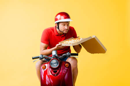 delivery man in helmet riding red scooter and smelling pizza in box on yellowの写真素材