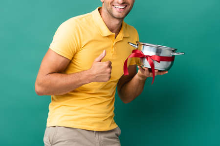cropped view of cheerful delivery man holding saucepan with ribbon and showing thumb up on blueの写真素材