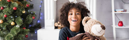 excited african american girl looking at camera while holding teddy bear on blurred background, bannerの写真素材