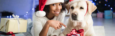 happy african american girl pointing with finger at gift box while lying on floor near labrador dog on blurred background, bannerの写真素材