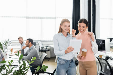 Smiling businesswomen showing yeah while holding digital tablet and coffee to go in officeの写真素材