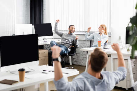 Multiethnic businesspeople showing yeah gesture while working near computers in officeの写真素材