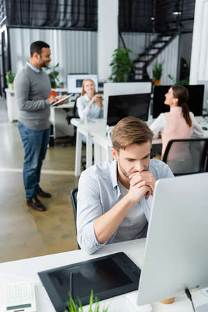 Pensive businessman looking at computer near graphics tablet and multicultural colleagues on blurred...