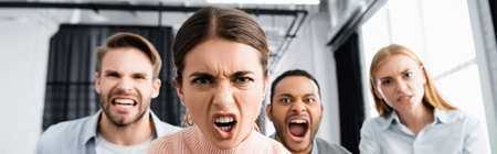 Angry businesswoman looking at camera near multiethnic colleagues on blurred background in office, bannerの写真素材