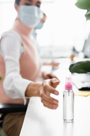 Businesswoman in medical mask pulling hand to sanitizer on table on blurred backgroundの写真素材