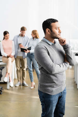Thoughtful indian businessman standing near colleagues working on blurred background in office