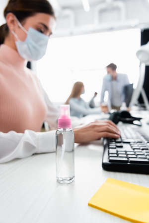 Bottle of hand sanitizer near businesswoman in medical mask using computer keyboard on blurred backgroundの写真素材