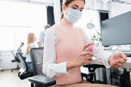 Businesswoman in medical mask using hand sanitizer in officeの写真素材