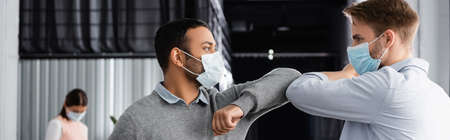 Multiethnic businessmen in medical masks giving high five with elbows in office, banner