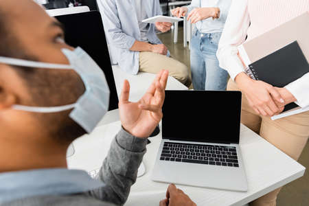 Businesspeople with digital tablet and notebook standing near indian colleague in medical mask in officeの写真素材