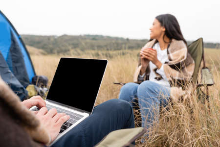 Man using laptop near african american girlfriend with cup on blurred background during camping in fieldの写真素材