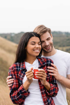 Smiling man with closed eyes hugging african american girlfriend with cup outdoors