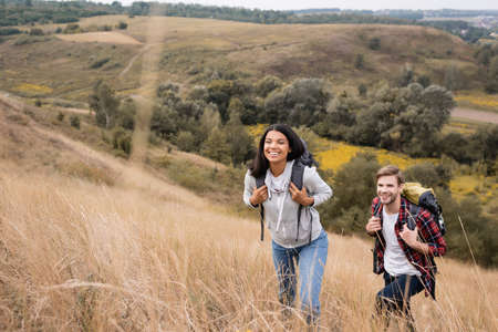 Interracial couple with backpacks walking on hill with grass during tripの写真素材