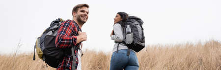 Smiling multiethnic hikers with backpacks walking on hill with sky at background, banner