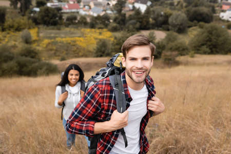 Cheerful traveler looking at camera near african american girlfriend on blurred background during tripの写真素材