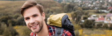 Smiling tourist with backpack looking at camera with landscape on blurred background, banner