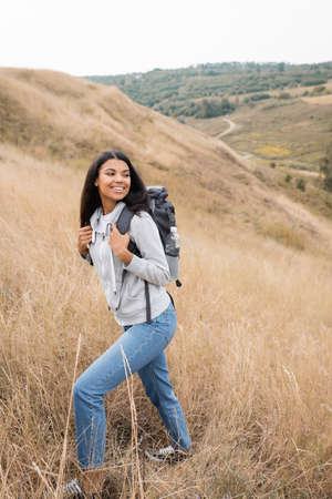 Positive african american hiker holding backpack while walking on hill with grassの写真素材
