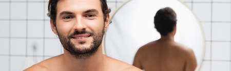 Bearded and shirtless man smiling at camera near mirror on blurred background in bathroom, bannerの写真素材